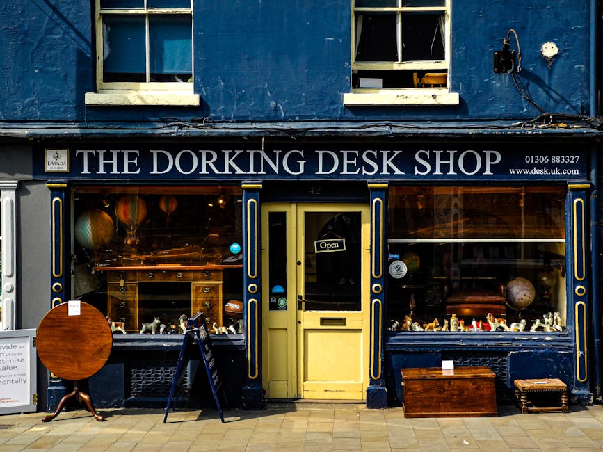 The storefront of The Dorking Desk Shop located on a street with a dark blue exterior and white trim around the windows and door. The shop's name is displayed on a black sign with white lettering abov