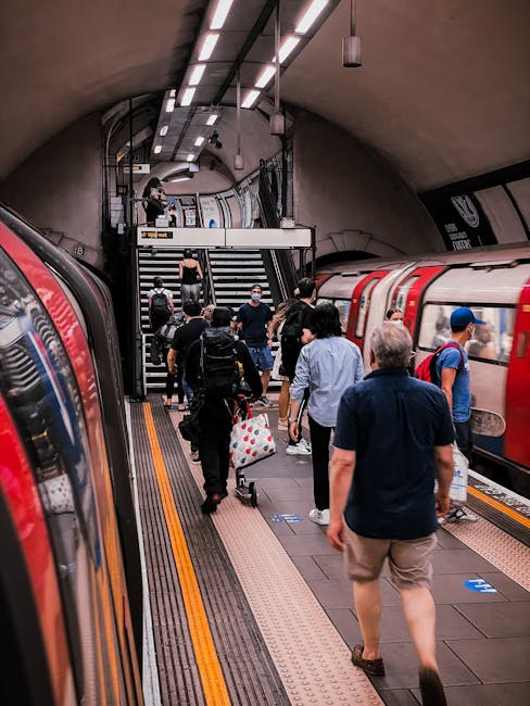 Inside a busy underground train station, several passengers are walking along the platform, some carrying luggage or backpacks. The platform has a tactile paving strip near the edge for safety, with a modern lighting system illuminating the curved ceiling. Two train carriages are visible on either side of the platform, their doors open for boarding. Passengers are seen ascending a staircase at the end of the platform, some wearing face masks. The scene captures the typical environment of a public transportation hub used during home relocation or moving logistics, with people preparing to board trains as part of their journey, supported by efficient transfer services like those offered by Man With a Van Bexleyheath.