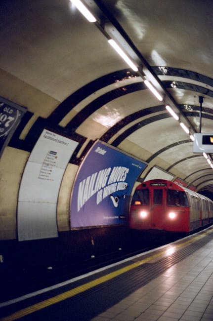 Inside a busy underground train station, several passengers are walking along the platform, some carrying luggage or backpacks. The platform has a tactile paving strip near the edge for safety, with a modern lighting system illuminating the curved ceiling. Two train carriages are visible on either side of the platform, their doors open for boarding. Passengers are seen ascending a staircase at the end of the platform, some wearing face masks. The scene captures the typical environment of a public transportation hub used during home relocation or moving logistics, with people preparing to board trains as part of their journey, supported by efficient transfer services like those offered by Man With a Van Bexleyheath.