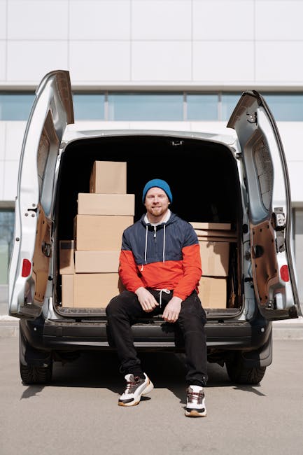 A young man with a beard, wearing a black cap and a white polo shirt, stands inside the open rear doors of a moving van loaded with stacked brown cardboard boxes, some wrapped in plastic film. The bac