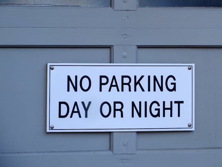 A close-up view of a rectangular traffic sign mounted on a metal pole against a bright blue sky. The sign displays a red circle with a diagonal line crossing out the letter 'P', indicating no parking,