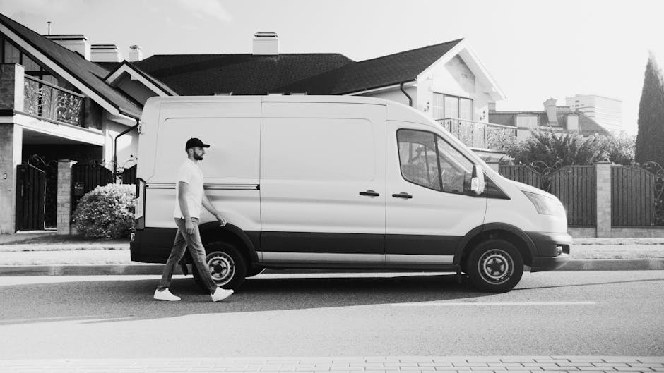 A man wearing a white t-shirt, dark jeans, white sneakers, and a black cap is walking alongside a white delivery van parked on a residential street in Bexleyheath. The van is positioned parallel to th